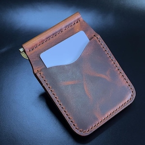 Puede incluir: Cartera de cuero marrón con clip para billetes. La cartera tiene costuras visibles y una tarjeta dentro. El clip para billetes es dorado. El cuero tiene un aspecto envejecido, lo que añade carácter al diseño.