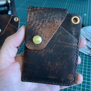 May include: A handcrafted, distressed leather wallet in shades of brown and black. The wallet features a flap closure with a brass button and a contrasting lighter brown accent. A stack of US dollar bills is visible in the background.