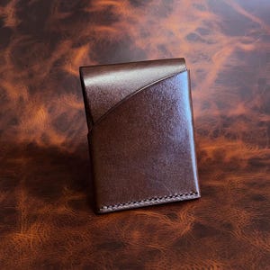 May include: A brown leather wallet with a diagonal design on the front. The wallet is rectangular and appears to be made of high-quality leather. The stitching is visible along the edges. The wallet is on a brown, textured surface.