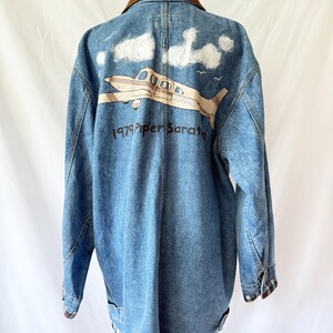 May include: Blue denim jacket with a brown corduroy collar. The back features a graphic of a plane with the text "1979 Piper Saratoga" and white cloud accents. The jacket has a checkered pattern trim.