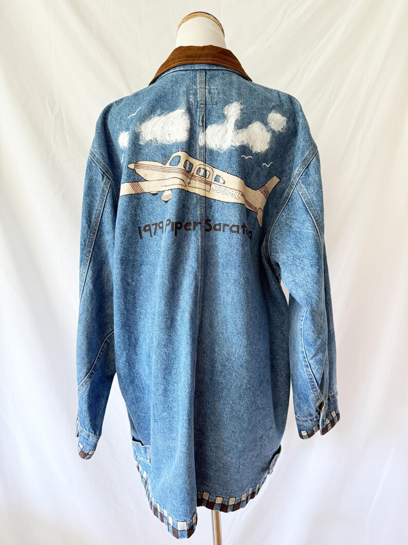 May include: Blue denim jacket with a brown corduroy collar. The back features a graphic of a plane with the text "1979 Piper Saratoga" and white cloud accents. The jacket has a checkered pattern trim.