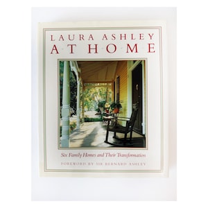 Puede incluir: Un libro titulado "LAURA ASHLEY AT HOME" con una fotografía de un porche con una mecedora. La portada tiene un fondo crema con texto rojo y un borde rojo. El subtítulo dice "Six Family Homes and Their Transformation".