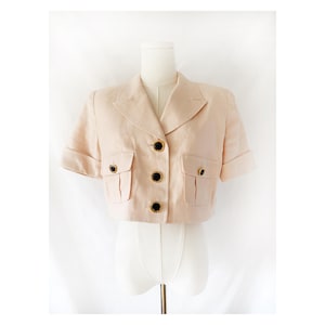 May include: A light peach-colored cropped blazer with short sleeves, two front pockets, and black buttons with gold rims. The blazer is displayed on a white mannequin against a white background. The jacket has a classic collar.