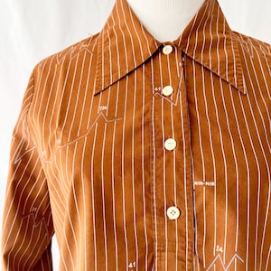 Può includere: Una camicia a maniche lunghe a righe marroni e bianche con colletto abbottonato. La camicia presenta una stampa grafica bianca di linee e numeri. Il testo "NIK-NIK" è stampato sulla camicia.