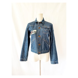 May include: A white ceramic mug with a black handle and the text "But First, Coffee" in a bold, black font. The mug is set against a blurred background, suggesting a cozy setting.