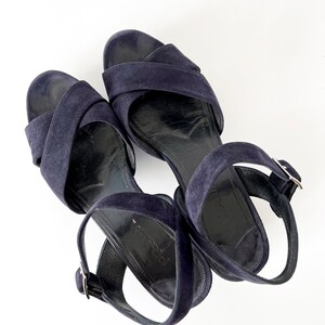 Op de afbeelding: Een paar donkerblauwe su&egrave;de sandalen met enkelbandjes en gespen.