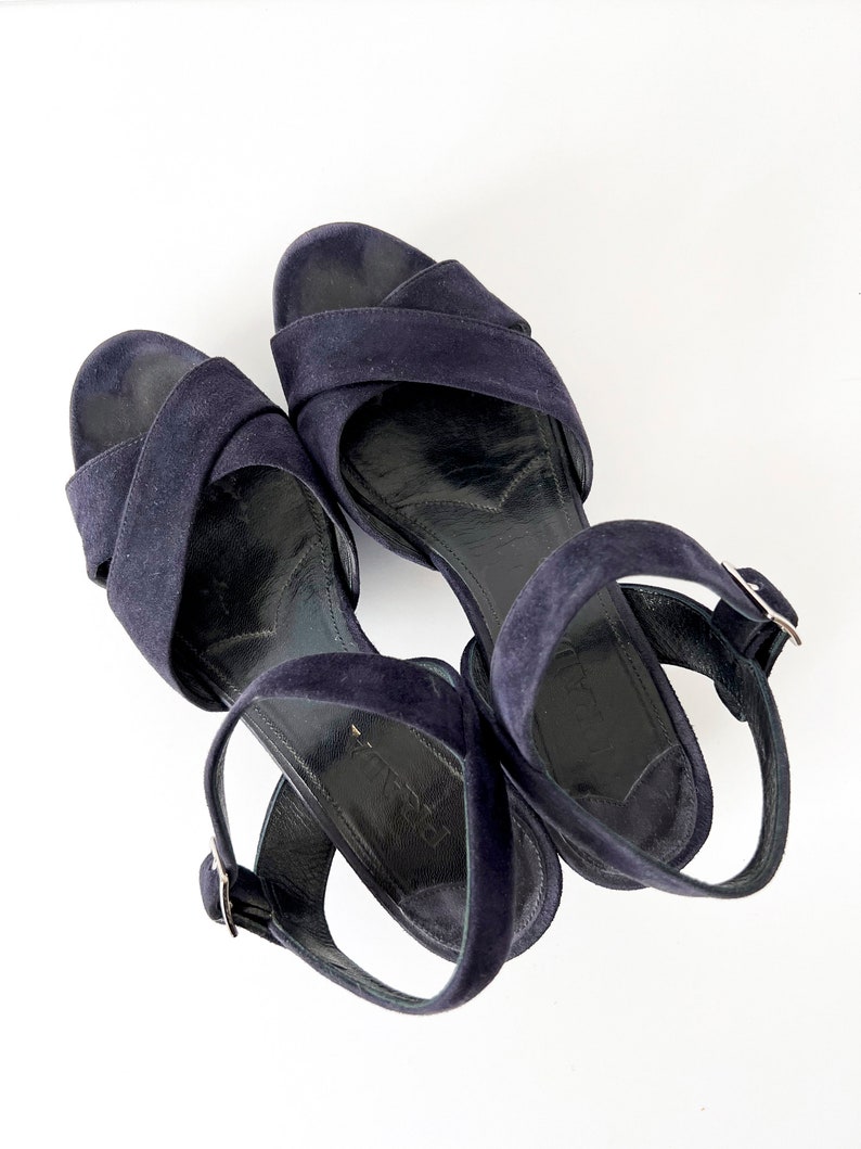 Op de afbeelding: Een paar donkerblauwe su&egrave;de sandalen met enkelbandjes en gespen.