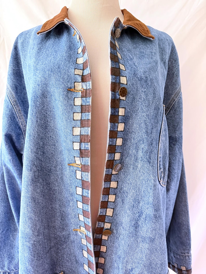 May include: Light blue denim jacket with a brown corduroy collar. The jacket features a checkered pattern in brown and white along the front edges and gold-colored buttons. A classic denim jacket.