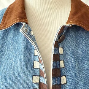 May include: Close-up of a light blue denim jacket with a brown suede collar. The jacket features a decorative pattern of brown and white squares along the button line. The jacket is buttoned up.
