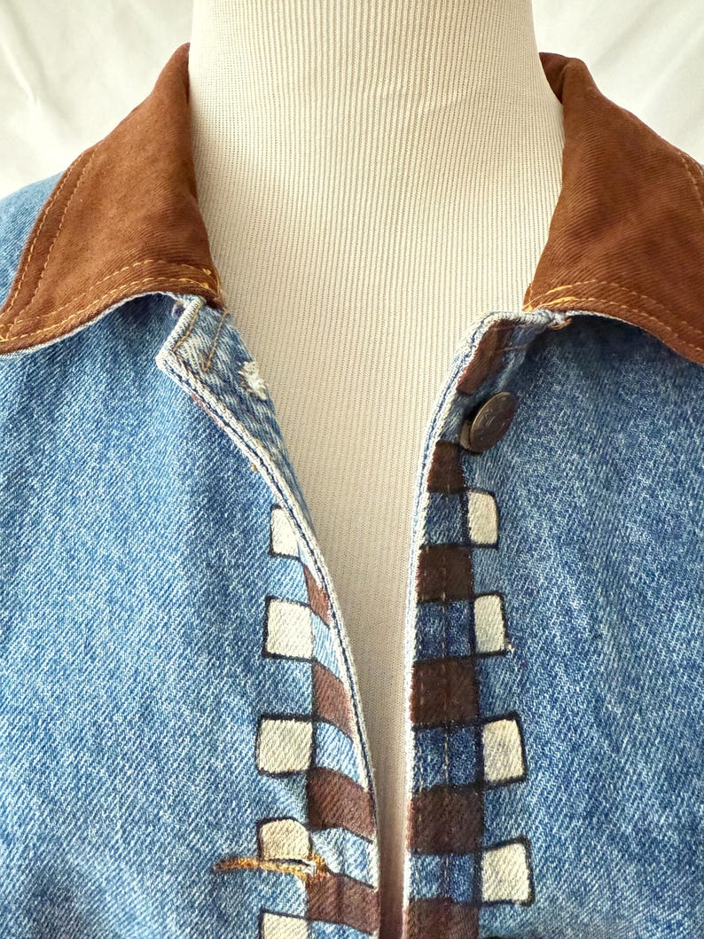 May include: Close-up of a light blue denim jacket with a brown suede collar. The jacket features a decorative pattern of brown and white squares along the button line. The jacket is buttoned up.