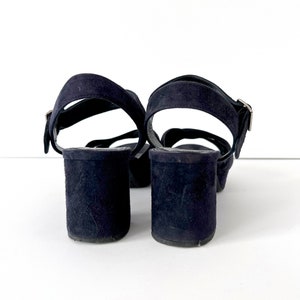 Op de afbeelding: Een paar donkerblauwe su&egrave;de sandalen met enkelbandjes en gespen.