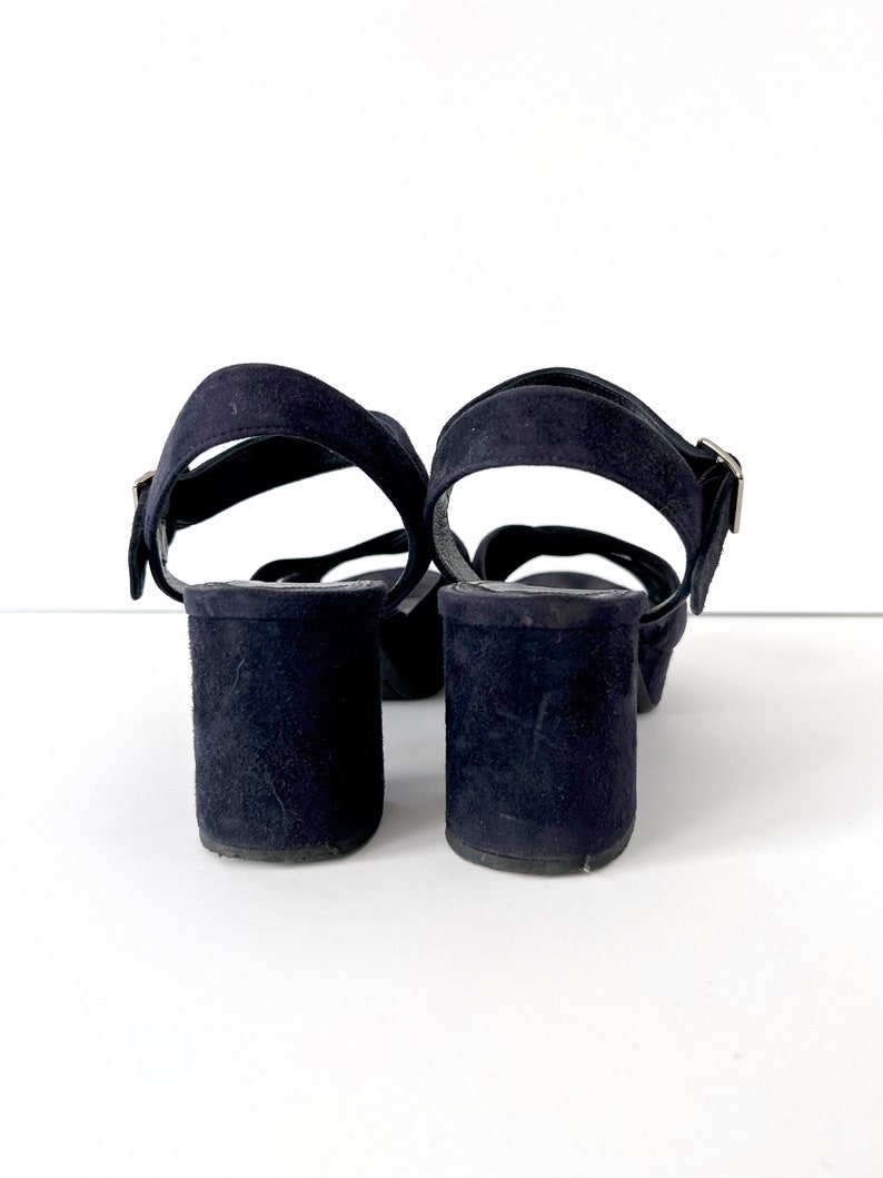 Op de afbeelding: Een paar donkerblauwe su&egrave;de sandalen met enkelbandjes en gespen.
