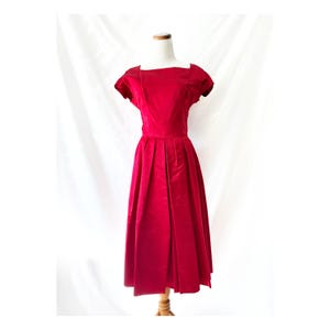 May include: A vintage red velvet dress with a square neckline and short sleeves. The dress features a fitted bodice and a pleated, midi-length skirt. The dress is displayed on a mannequin against a white backdrop.