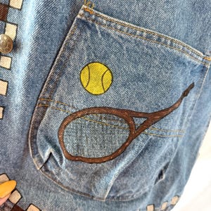May include: Close-up of a light blue denim vest with a pocket featuring a yellow tennis ball and a brown tennis racket design. The vest has a brown and white checkered pattern along the edges and a silver button.