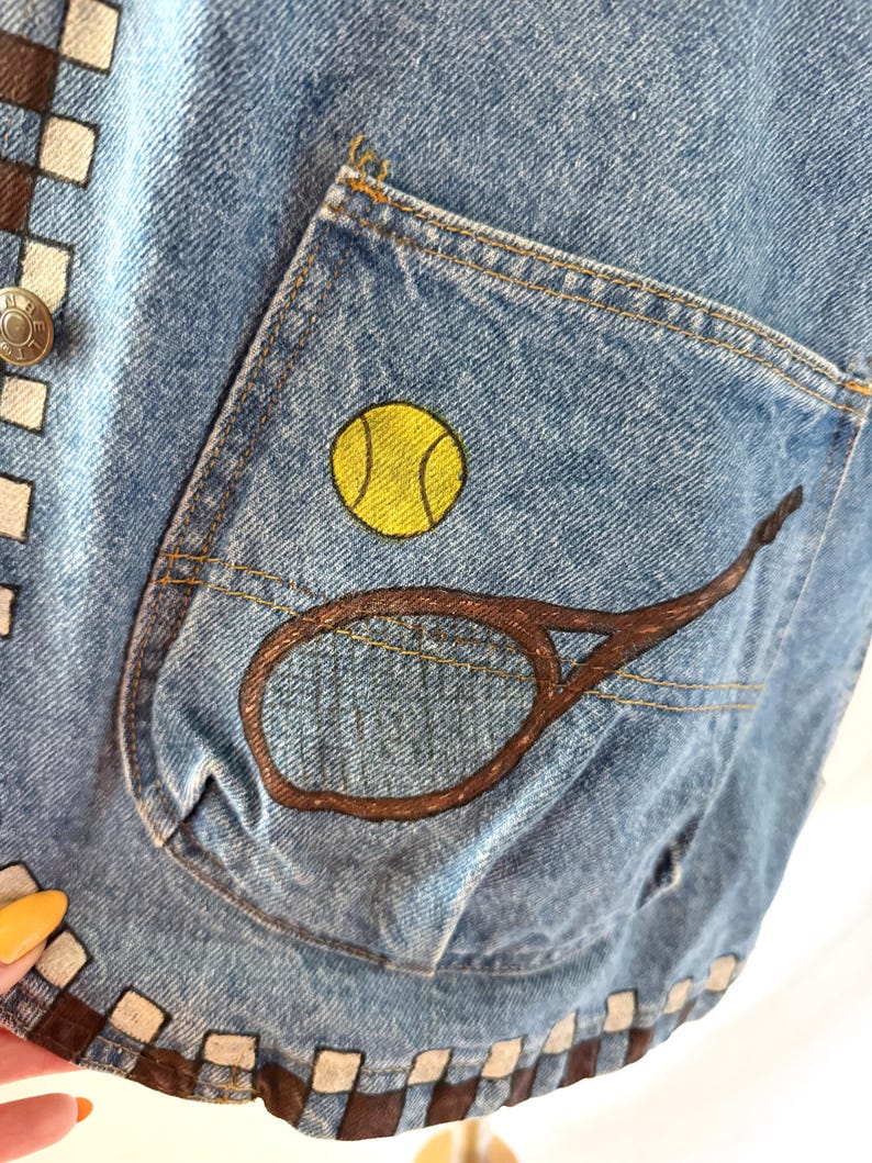 May include: Close-up of a light blue denim vest with a pocket featuring a yellow tennis ball and a brown tennis racket design. The vest has a brown and white checkered pattern along the edges and a silver button.