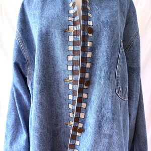 May include: A light blue denim jacket with a brown corduroy collar. The jacket features a checkered pattern in brown and white along the front and sleeve cuffs. It has large pockets and brass-colored buttons.