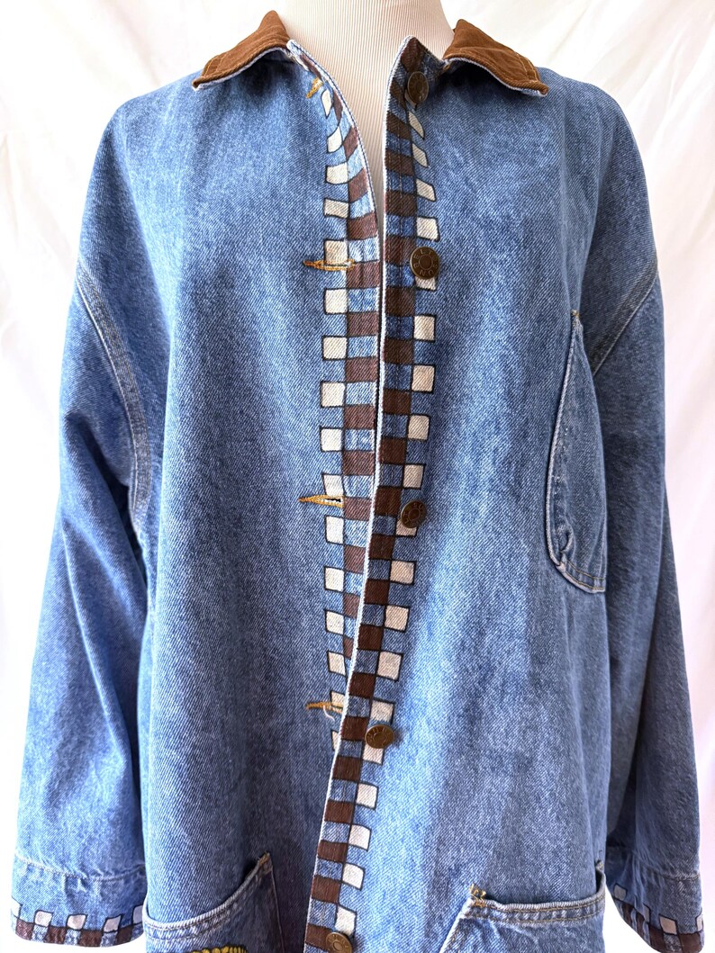 May include: A light blue denim jacket with a brown corduroy collar. The jacket features a checkered pattern in brown and white along the front and sleeve cuffs. It has large pockets and brass-colored buttons.