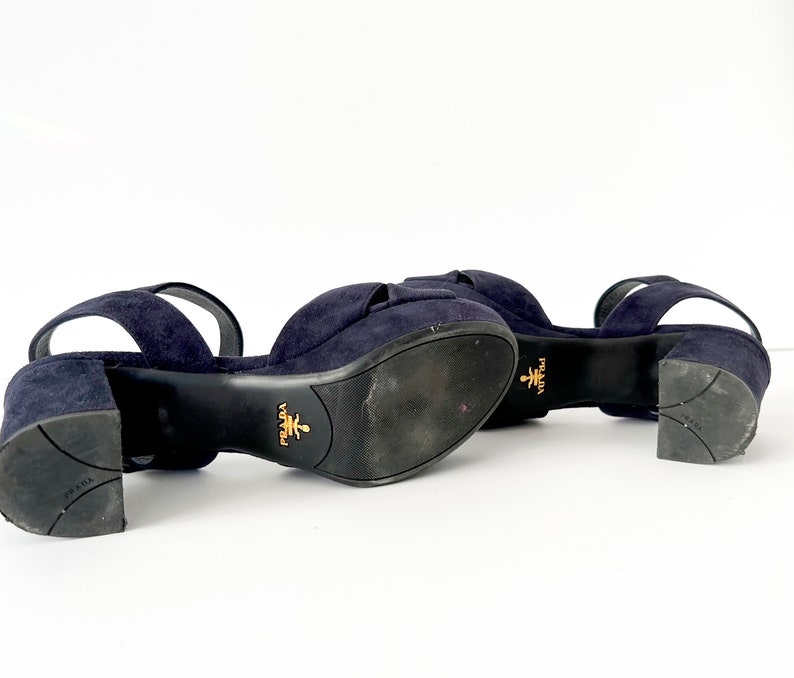 Op de afbeelding: Een paar donkerblauwe su&egrave;de sandalen met enkelbandjes. De sandalen hebben een blokhak en de merknaam "Prada" is zichtbaar op de zool.