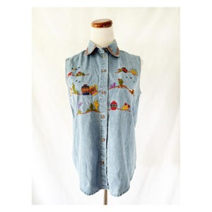 May include: A light blue sleeveless denim shirt featuring a button-down front and a pointed collar. The shirt is decorated with colorful embroidered motifs of desert landscapes, including cacti, pottery, and suns, scattered across the front panels.