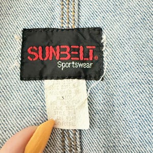 May include: Close-up of a denim jacket with a brown leather collar and seams. The black label reads "SUNBELT Sportswear" in red lettering. A white tag below indicates the size is "LARGE".