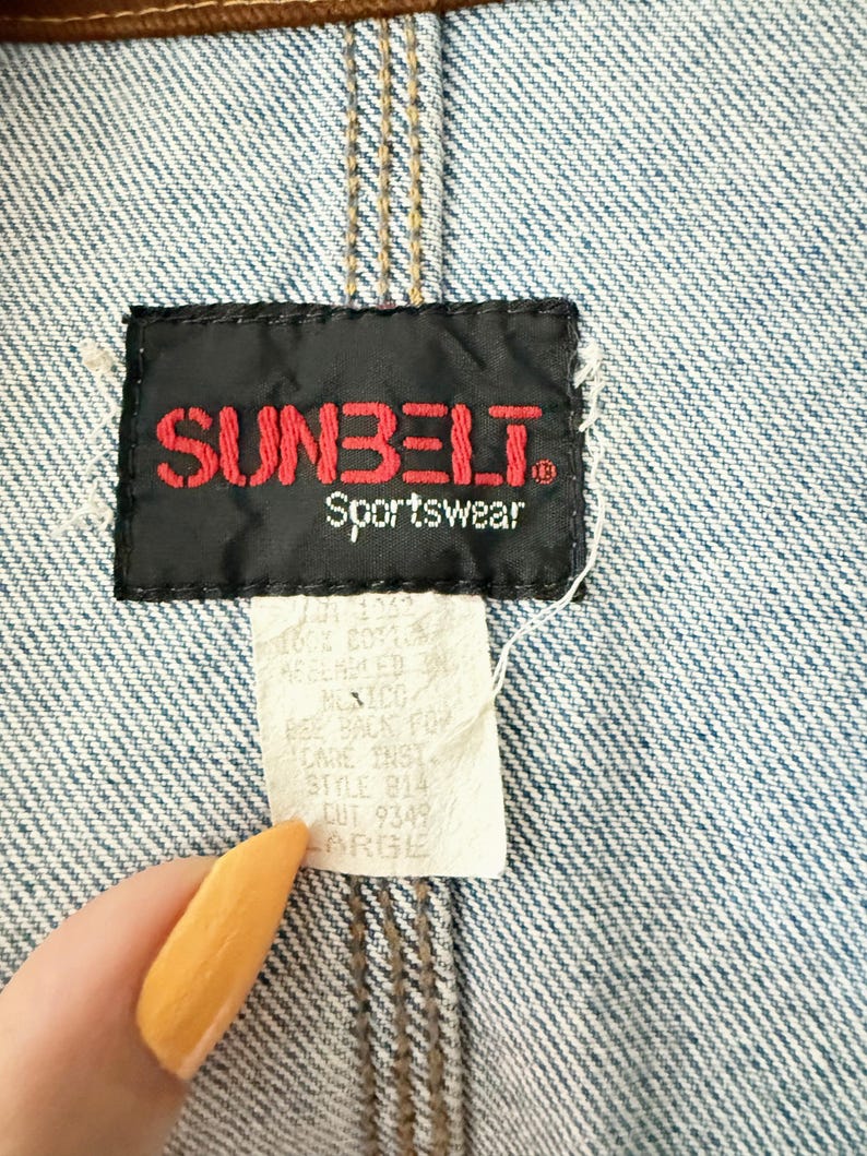 May include: Close-up of a denim jacket with a brown leather collar and seams. The black label reads "SUNBELT Sportswear" in red lettering. A white tag below indicates the size is "LARGE".