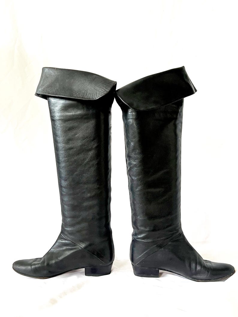 May include: A pair of black leather over-the-knee boots with a folded-over cuff at the top. The boots have a low heel and a pointed toe.