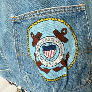 May include: Light blue denim jacket with a pocket featuring a United States Coast Guard patch. The patch includes an anchor, the words "United States Coast Guard," and "Semper Paratus 1790." The jacket has a brown and white trim.