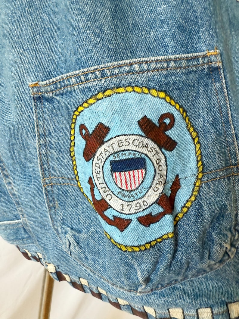 May include: Light blue denim jacket with a pocket featuring a United States Coast Guard patch. The patch includes an anchor, the words "United States Coast Guard," and "Semper Paratus 1790." The jacket has a brown and white trim.