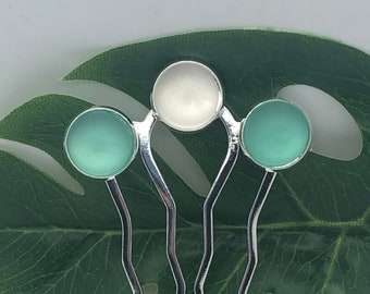 May include: Three silver hair combs with decorative round turquoise and white glass accents. The combs are on a green leaf.
