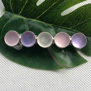May include: A silver hair clip with five round, translucent stones. The stones are in shades of purple, pink, and white.