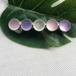 May include: A silver hair clip with five round, iridescent stones. The stones are in shades of pink, purple, and white.