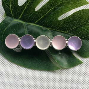 May include: A silver hair clip with five round, translucent stones. The stones are in shades of pink, purple, and white.