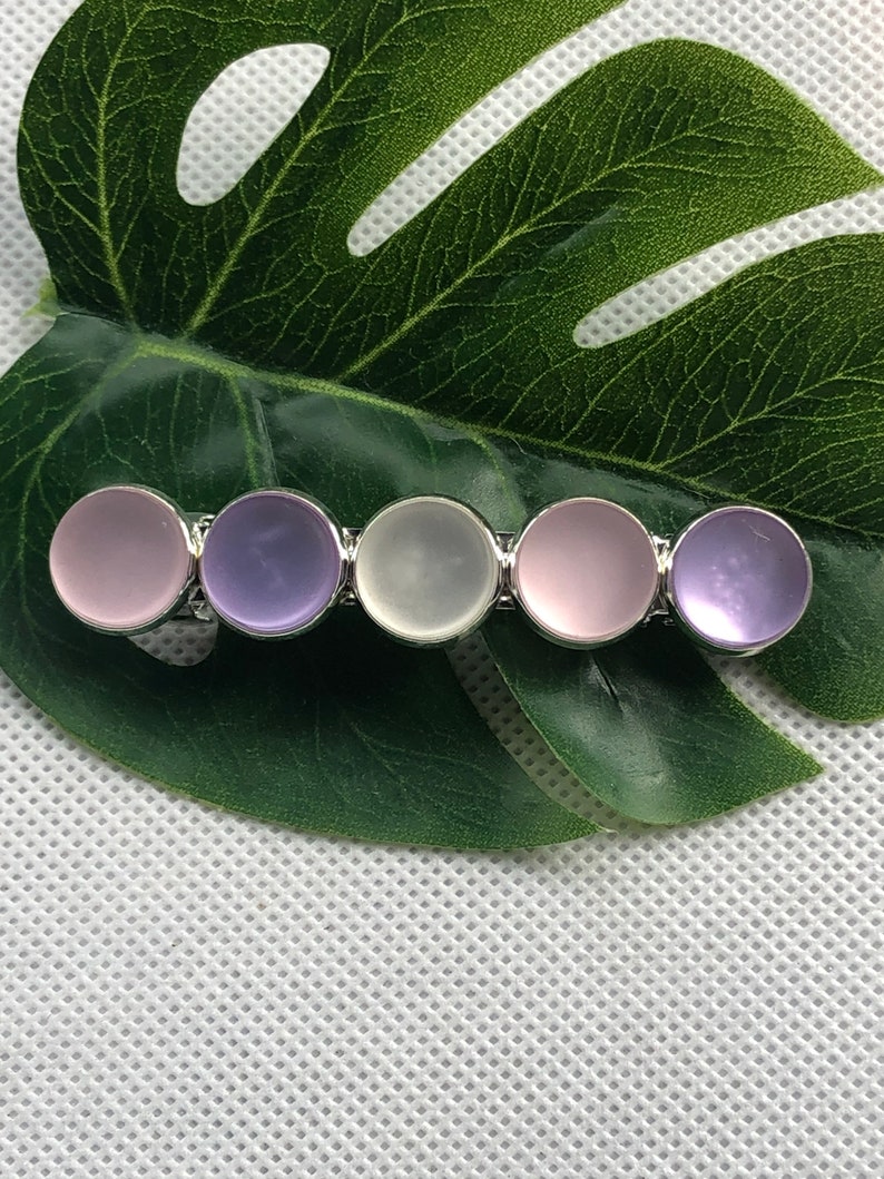 May include: A silver hair clip with five round, translucent stones. The stones are in shades of pink, purple, and white.