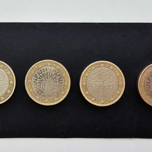 Puede incluir: Cuatro monedas de euro de color plateado con un diseño de árbol y el año 2002 impreso en ellas. Las monedas están dispuestas en fila sobre una superficie negra.