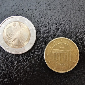 May include: Two Euro coins on a black surface. One coin is silver and gold, featuring an eagle and the year 2002. The other coin is gold, depicting a building and the year 2002. Both coins have stars around the edges.