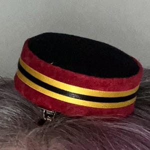 May include: A small, decorative hat in a pillbox style. The hat features a black top and a red band with two yellow stripes. A silver clip is attached to the bottom. The hat is likely a hair accessory.