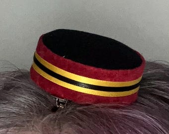 Può includere: Un piccolo cappello decorativo in stile pillbox. Il cappello presenta una parte superiore nera e una fascia rossa con due strisce gialle. Una clip argentata è attaccata in basso. Il cappello è probabilmente un accessorio per capelli.