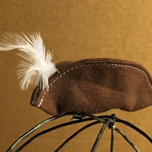 May include: A brown felt hat with a white feather attached to the side. The hat is sitting on a wire frame.