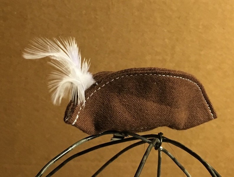 May include: A brown felt hat with a white feather attached to the side. The hat is sitting on a wire frame.