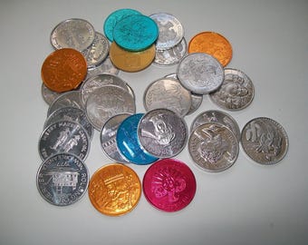 Könnte beinhalten: Eine Sammlung verschiedener Münzen in Silber, Orange, Türkis, Gold und Pink. Einige Münzen haben geprägte Designs, andere Text. Die Münzen sind auf einer weißen Oberfläche verstreut.