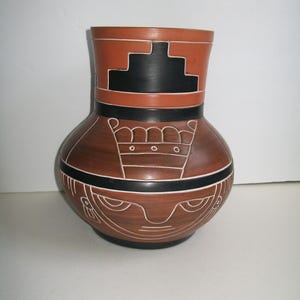 May include: A brown ceramic vase with a wide base and a narrow neck. The vase features geometric designs in black and white, including a stepped pattern at the top and stylized faces around the base. The vase is approximately 10 inches tall.