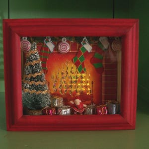 May include: A red-framed shadow box featuring a miniature Christmas scene with a fireplace backdrop, hanging stockings, peppermint candies, a small flocked pine tree, wrapped gifts, and a tiny Santa Claus figure standing in the center.