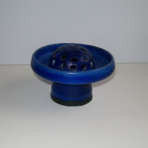 May include: A cobalt blue ceramic flower frog featuring a wide, circular bowl-shaped top with a central, perforated dome. The base is cylindrical and dark, providing a stable foundation for this functional floral arrangement tool.