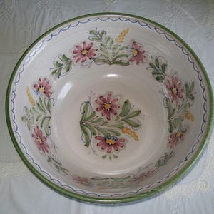 May include: A large, white ceramic bowl with a floral design. The bowl features pink flowers, green leaves, and yellow accents. The rim has a green border with a scalloped blue edge. This decorative bowl is ideal for serving.