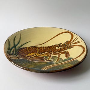 May include: A ceramic plate with a hand-painted lobster design. The plate features a cream-colored background with a brown lobster, green seaweed, and blue water details. The rim is a darker brown.