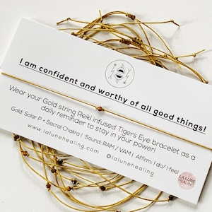 May include: Gold string bracelet with tiger's eye beads. The bracelet is on a white card with the text "I am confident and worthy of all good things!" and "Wear your Gold string Reiki infused Tigers Eye bracelet as a daily reminder to stay in your power!"  Gold: Solar P + Sacral Chakra | Sound: RAM/VAM | Affirm: I do/ I feel www.lalunehealing.com @lalunehealing LA LUNE HEALING"