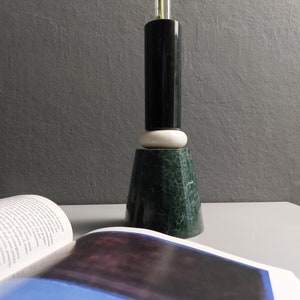 Könnte beinhalten: Eine schwarze und grüne Marmorvase mit einem weißen Marmorsockel. Die Vase ist mit einem einzigen grünen Stiel gefüllt.