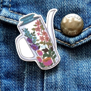 May include: A white ceramic teapot with a floral pattern in shades of pink, blue, purple, and green. The teapot has a spout and a handle. The teapot is on a blue denim background.