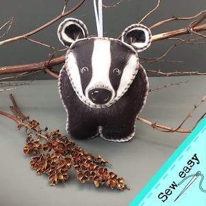 May include: A black and white felt badger ornament with a white stripe down its face. The ornament is hanging from a white string and is surrounded by brown branches. The text "Sew easy" is in the bottom right corner of the image.