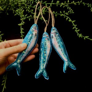 May include: Three blue and white felt fish ornaments with a painted design. Each fish has a loop of twine for hanging.